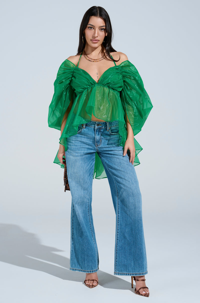 A woman faces forward against a light background, wearing the AN ELEGANT EVENING ORGANZA BLOUSE in sheer kelly green with off-the-shoulder voluminous sleeves, styled with blue wide-leg jeans, brown heeled sandals, and layered necklaces.