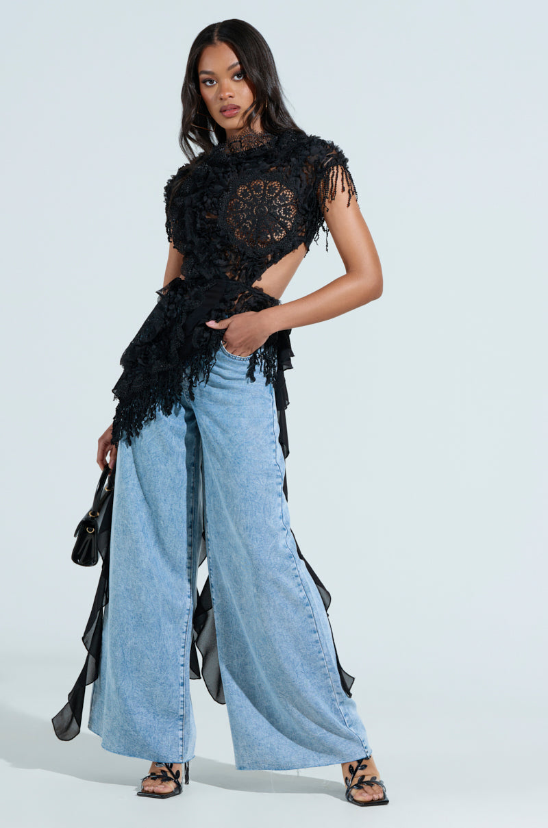 A woman poses against a light background wearing the THINK ABOUT ME TOP IN BLACK, featuring intricate floral lace and open sides, paired with wide-leg light blue jeans. She completes her look with a small black purse, strappy heels, hairstyle in loose waves, and a sleek metallic back zip for flair.