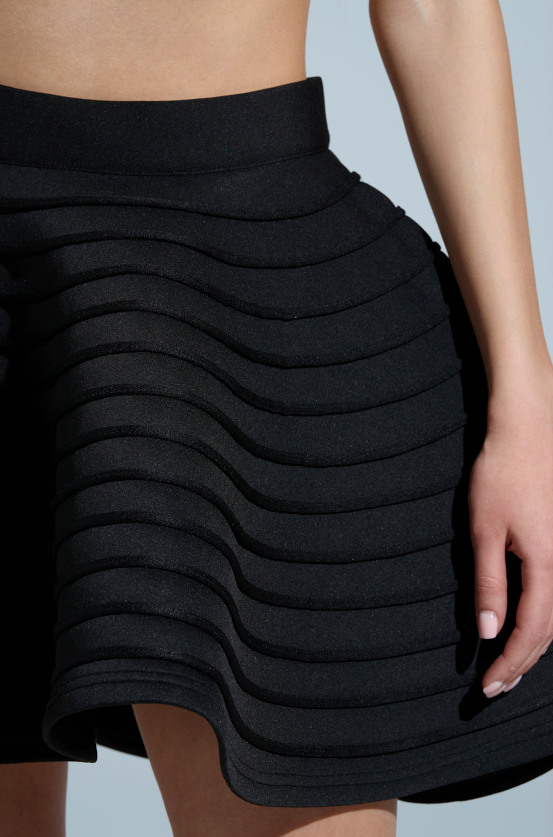 A person models the short, black DANCING QUEEN MINI SKIRT featuring layered, ruffled stripes creating a textured, wavy silhouette. Their hand rests gracefully at their side against a light gray background.