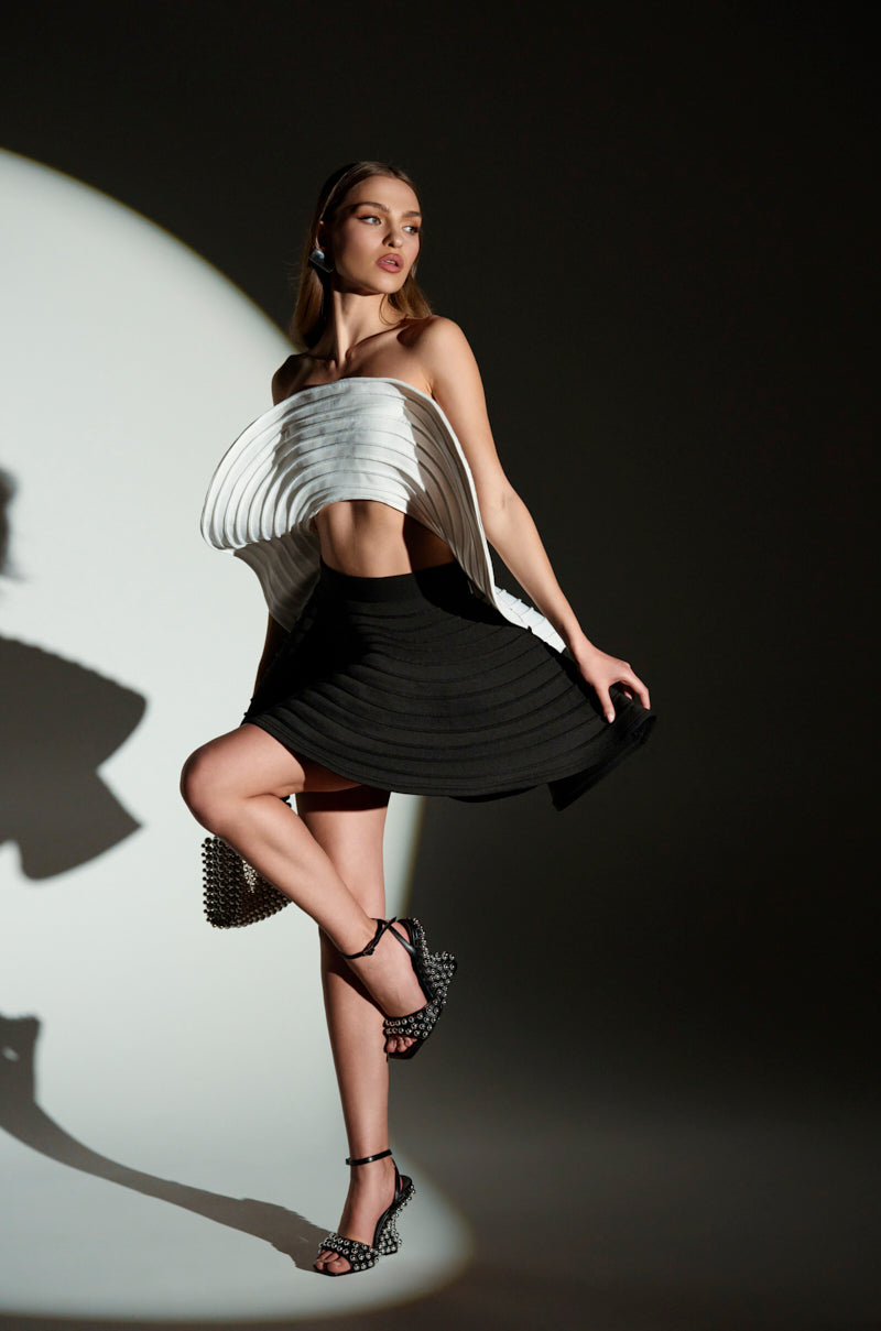 A model showcases a sculptural white layered top and the flared black Dancing Queen Mini Skirt. She pairs this with studded high heels, standing against dramatic lighting that casts her shadow on the wall.