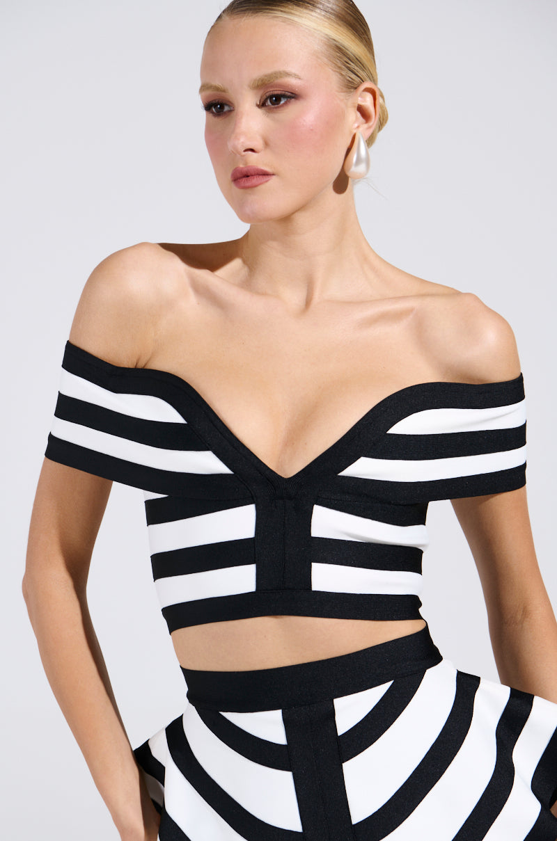 Modeling the WAITING FOR YOU STRIPE BLOUSE, an individual poses against a simple backdrop. The chic off-the-shoulder top features striking black-and-white stripes and patterns, complemented by slicked-back hair and large hoop earrings for an effortlessly stylish look.