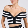 Modeling the WAITING FOR YOU STRIPE BLOUSE, an individual poses against a simple backdrop. The chic off-the-shoulder top features striking black-and-white stripes and patterns, complemented by slicked-back hair and large hoop earrings for an effortlessly stylish look.