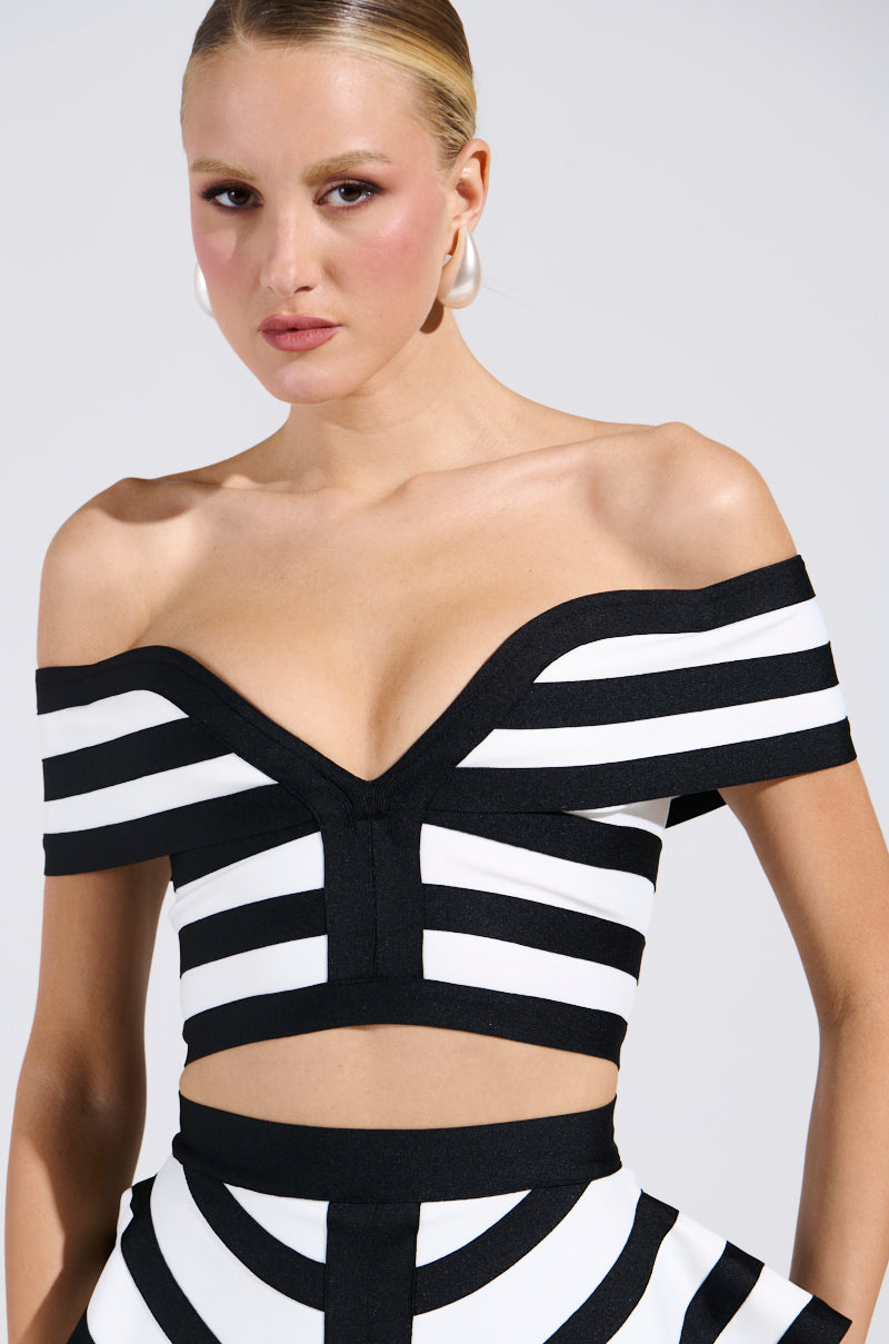 Wearing the WAITING FOR YOU STRIPE BLOUSE paired with a high-waisted skirt, this outfit's black-and-white off-the-shoulder geometric pattern complements short hair and hoop earrings, creating a chic statement against a plain white backdrop.