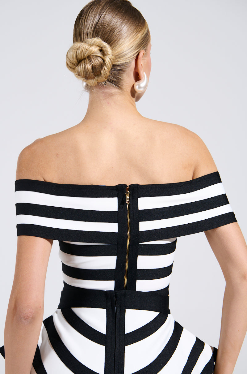 The rear view of a woman with blond hair in a bun showcases the WAITING FOR YOU STRIPE BLOUSE, an off-the-shoulder, black-and-white striped top with a central zipper and flared waist. She accessorizes with large pearl-like earrings against a simple backdrop.