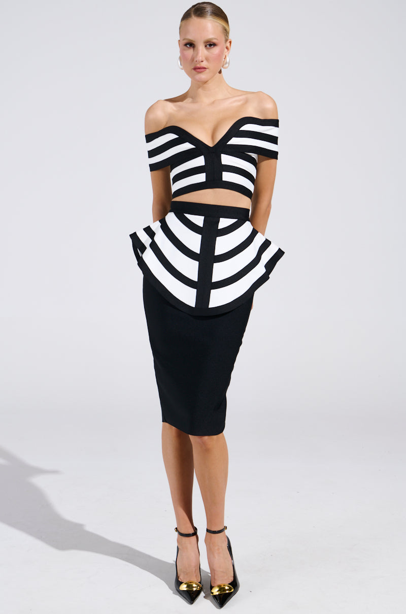 The individual wears the WAITING FOR YOU MIDI SKIRT, featuring a black and white off-the-shoulder dress with geometric patterns, a fitted top with cut-outs, and a bodycon silhouette. They pair it with black pointed shoes against a plain backdrop.