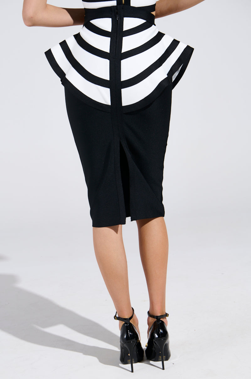 A person is wearing the WAITING FOR YOU MIDI SKIRT, a fitted black pencil skirt with a white and black striped peplum overlay. Paired with black high heels, the image taken from behind highlights the bodycon silhouette, back slit, and the person's legs.