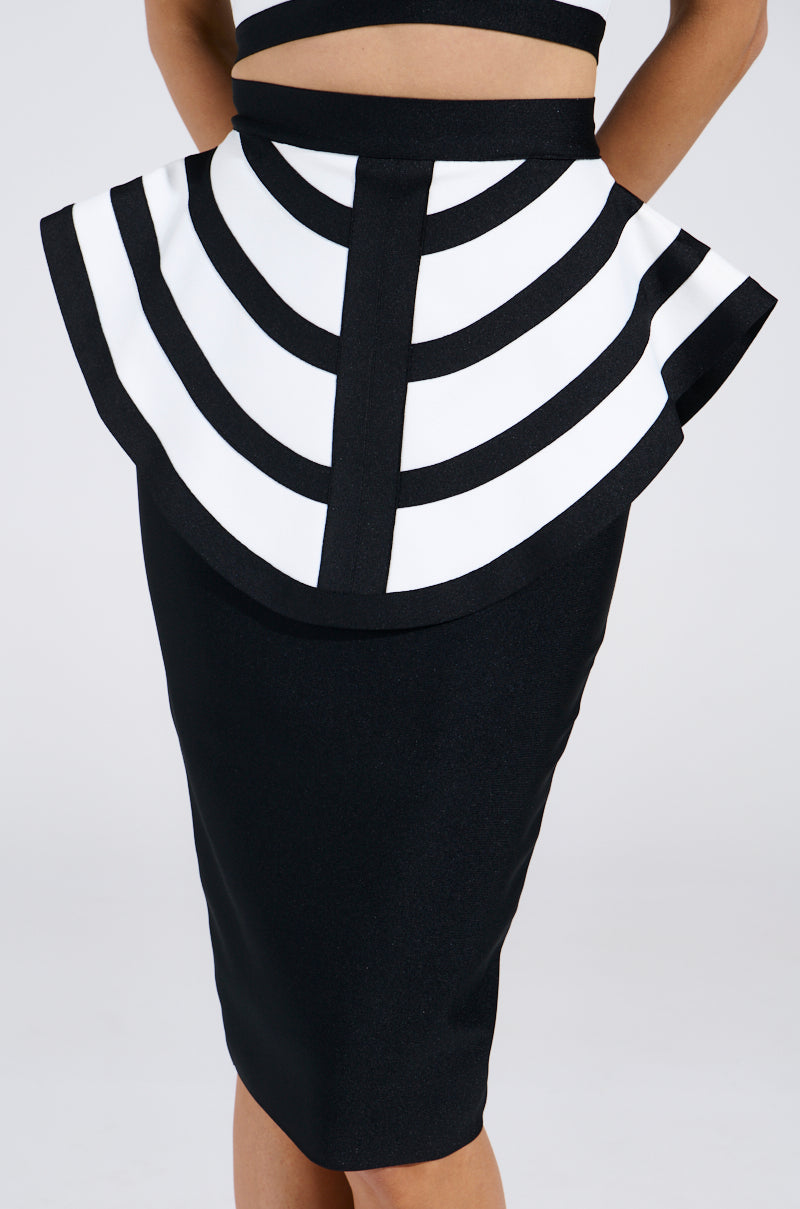 A person is wearing the WAITING FOR YOU MIDI SKIRT, featuring a structured flare with black and white geometric stripes at the waist. Their hands rest on their hips, highlighting its bold peplum overlay for a modern look.