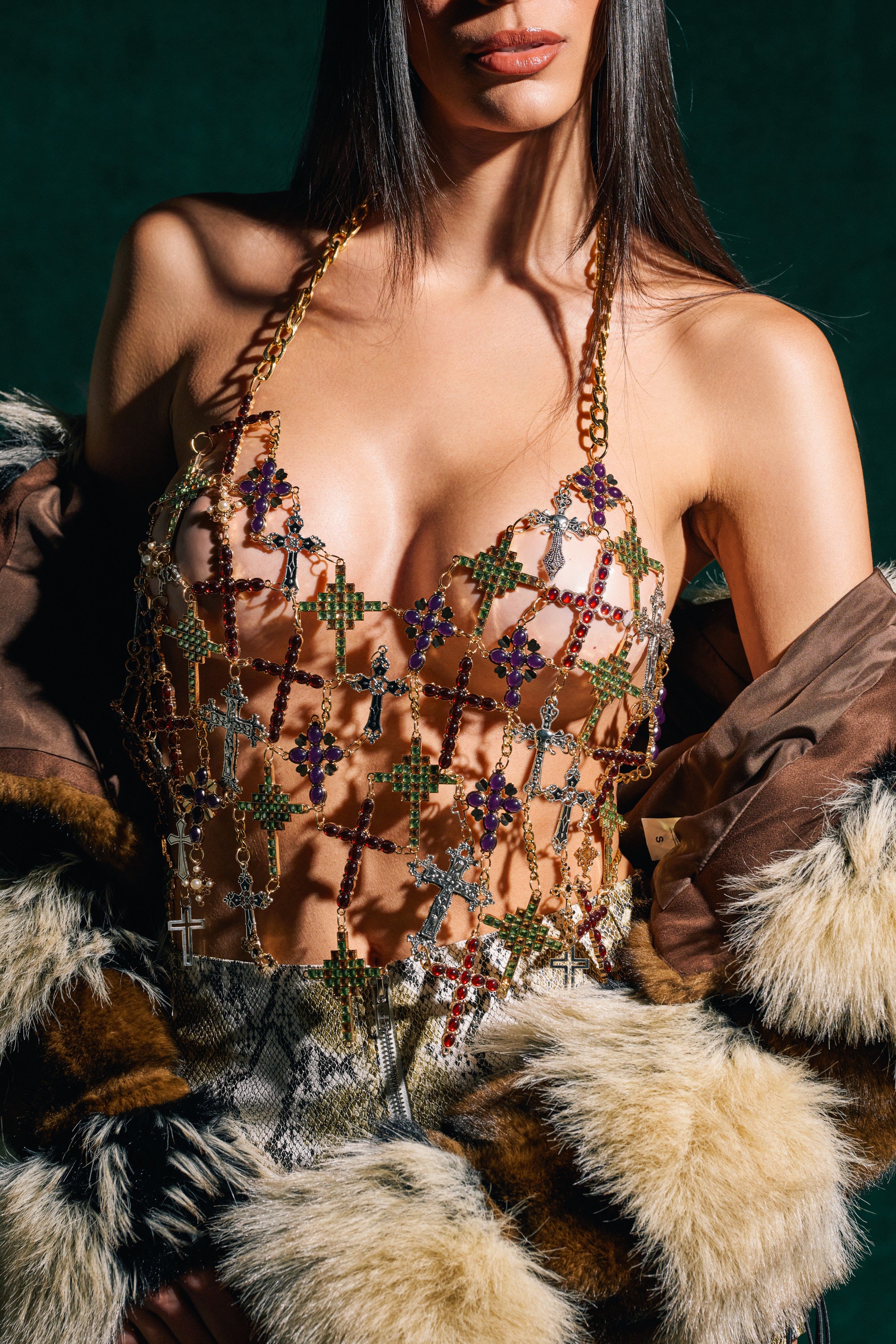 A woman wearing the BLESSED BRALETTE with colorful gems, a fur-trimmed coat draped over her arms, and snakeskin-patterned pants. Her long, straight hair is down, and her face is partially out of frame.