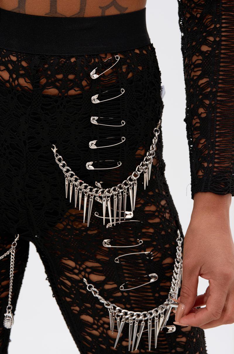 A close-up of a person wearing TRY ME LACE LEGGING—black, ripped leggings with silver safety pins, metal chains with spikes. Their hand rests by the chains, paired with a matching crocheted top.