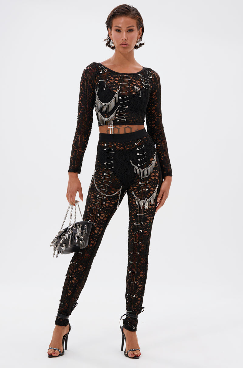 A woman stands confidently in the TRY ME LACE LONG SLEEVE WITH PINS jumpsuit, featuring black lace, cut-outs, sheer mesh top details, and silver pins. She carries a black handbag with metal accents and wears black strappy heels against a white background.