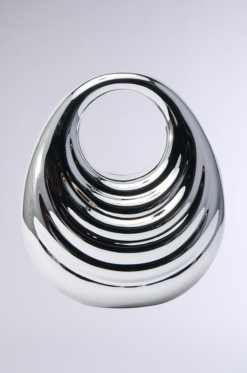 The STAY IN MY LANE ACRYLIC PURSE is a glossy, reflective metallic piece with a circular opening at the top. It boasts smooth, curved layers that resemble an avant-garde accessory's depth and reflection on its surface, all set against a simple light gray background.
