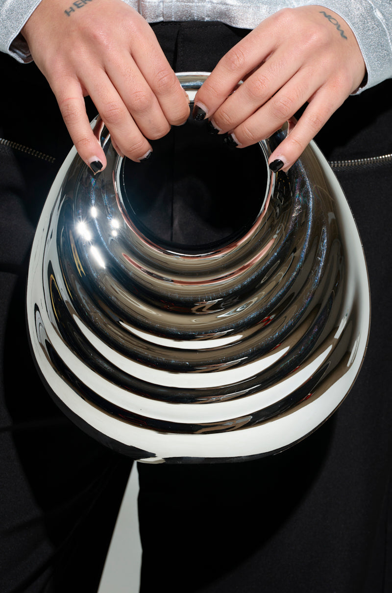A person showcases an avant-garde accessory, the STAY IN MY LANE ACRYLIC PURSE, in front of their waist. Sporting black nail polish and attired in a silver top with black pants, they create a striking ensemble accentuated by the purse’s reflective metallic design.