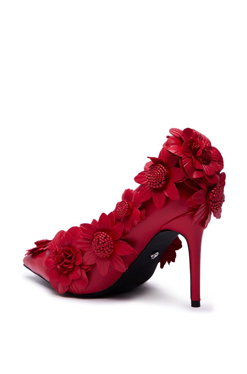 The AZALEA WANG ZARAH RED FLOWER PUMP features a striking stiletto heel, faux leather construction, and bold red flower appliqués decorating the heel, sides, and back—a standout floral design on a white background.