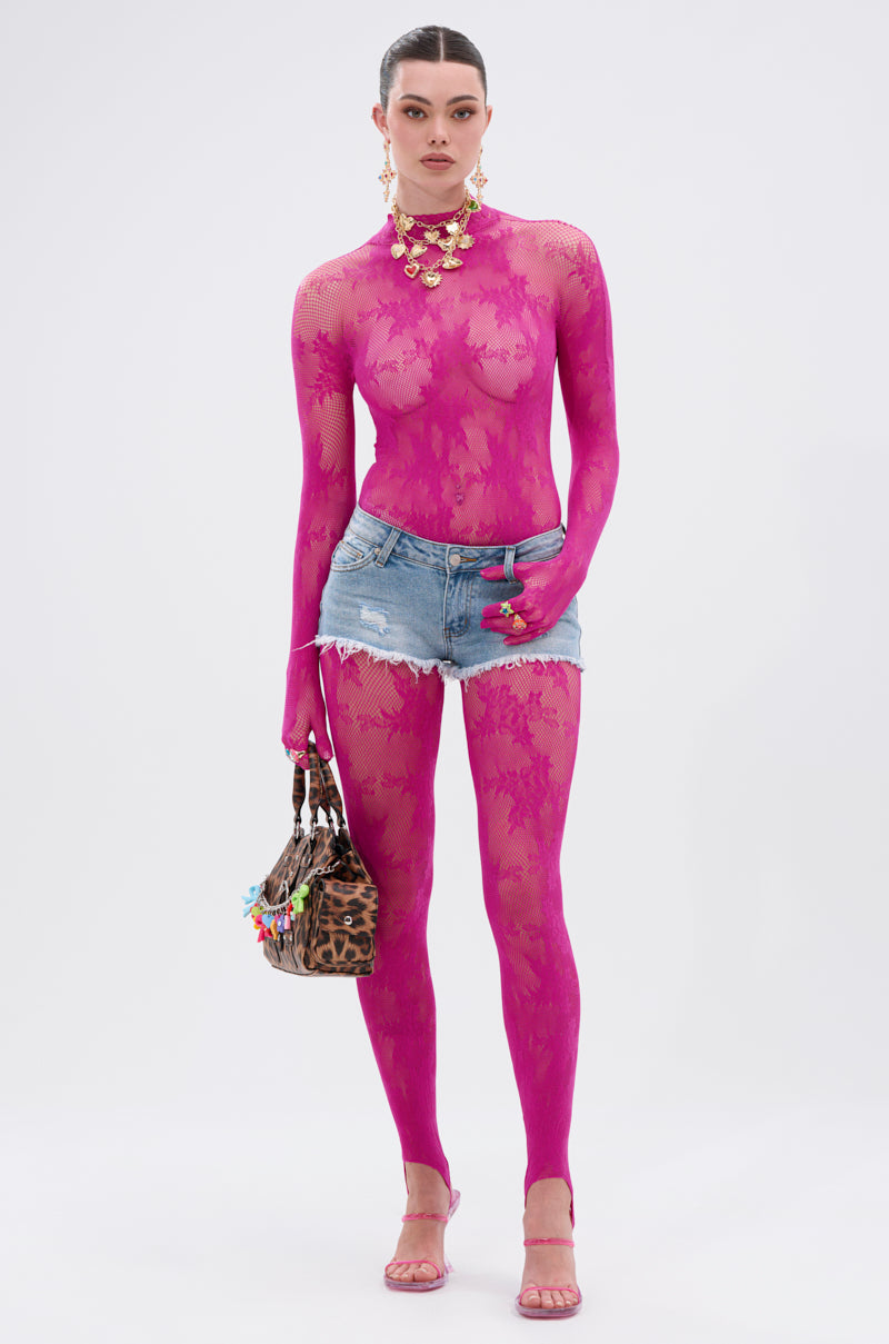 A woman stands against a white background wearing the MAIN MOMENT STRETCH LACE JUMPSUIT in sheer bright pink floral lace, styled with denim shorts, pink heels, statement jewelry, and a small leopard print handbag.