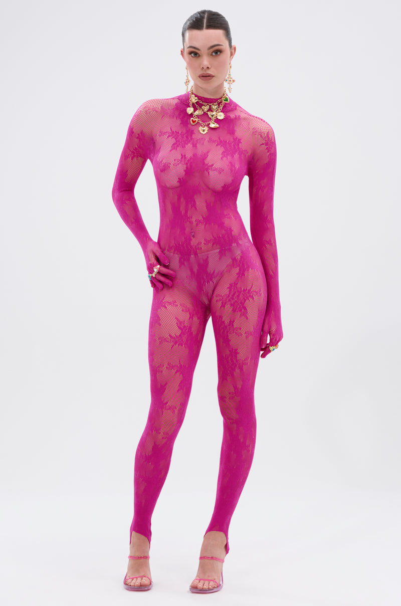A person poses confidently against a white background in the MAIN MOMENT STRETCH LACE JUMPSUIT, a sheer floral lace piece in bright pink with a mock neck, styled with matching heels, bold gold jewelry, and sleeked-back hair.