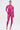 A person poses confidently against a white background in the MAIN MOMENT STRETCH LACE JUMPSUIT, a sheer floral lace piece in bright pink with a mock neck, styled with matching heels, bold gold jewelry, and sleeked-back hair.