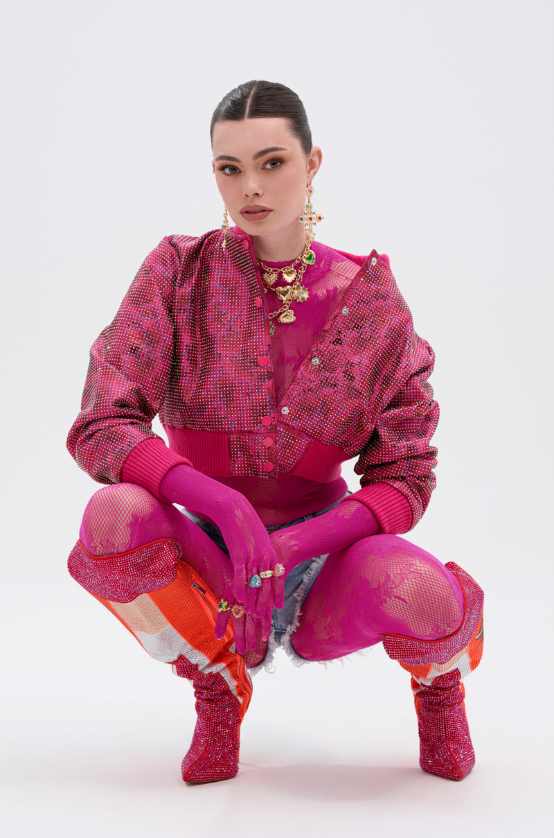 A woman squats against a white background, wearing the MAIN MOMENT STRETCH LACE JUMPSUIT with bright pink patterns, matching gloves, floral lace tights, layered boots, gold jewelry, and slicked-back hair as she looks confidently at the camera.