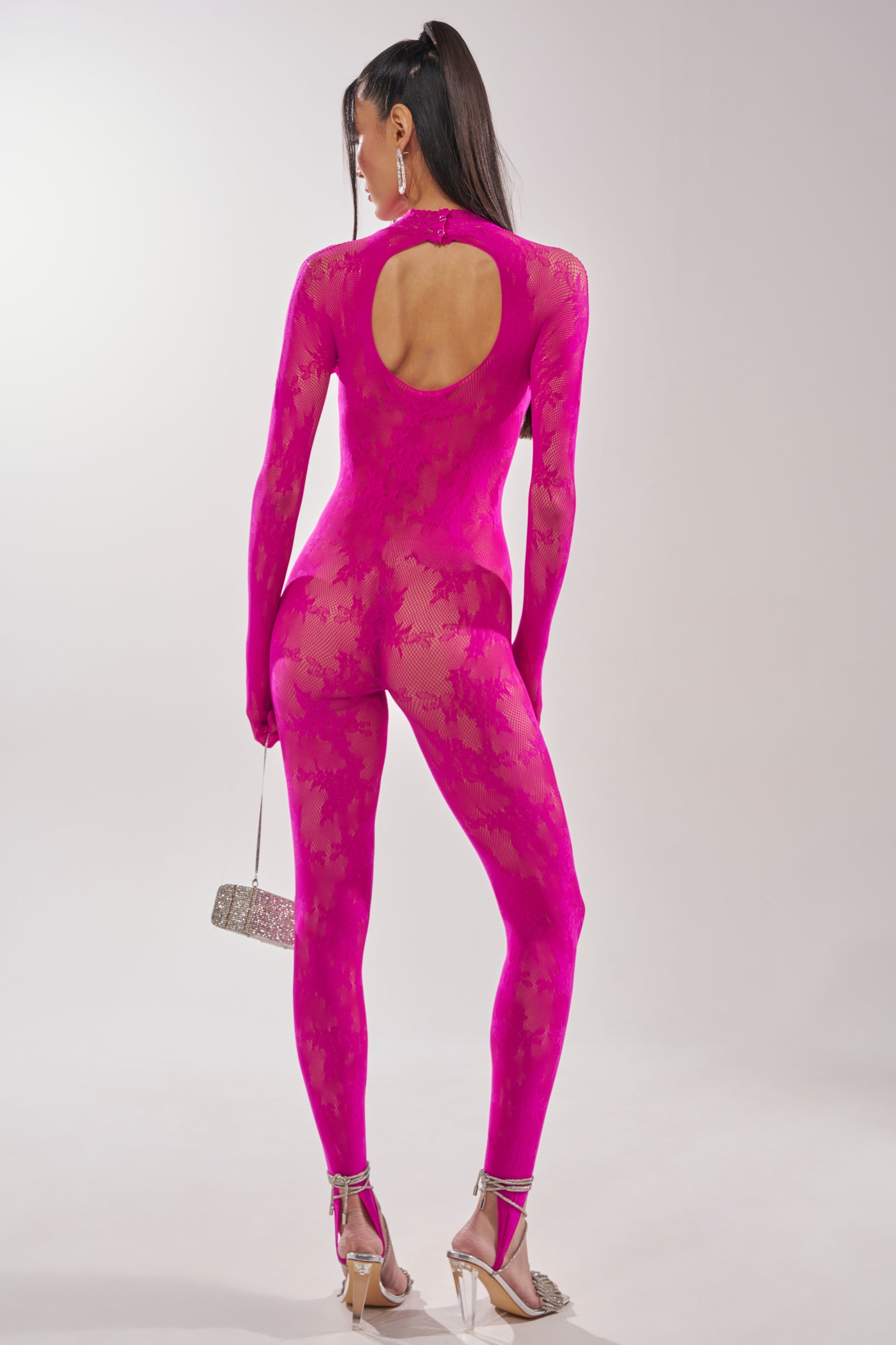 A person faces away, wearing the MAIN MOMENT STRETCH LACE JUMPSUIT in bright pink floral lace with a back keyhole cutout. They hold a small handbag and wear clear high-heeled sandals against a plain light background. 8