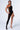 A person poses confidently in the ICONS ONLY STRETCH BODYSTOCKING, a stylish one-shoulder black piece with an asymmetrical silhouette and geometric cutouts along one side and leg. They wear transparent high-heeled shoes and flaunt a noticeable skull tattoo on their thigh against a plain, light-colored background.