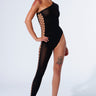 A person poses confidently in the ICONS ONLY STRETCH BODYSTOCKING, a stylish one-shoulder black piece with an asymmetrical silhouette and geometric cutouts along one side and leg. They wear transparent high-heeled shoes and flaunt a noticeable skull tattoo on their thigh against a plain, light-colored background.