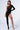 A person confidently poses in the ICONS ONLY STRETCH BODYSTOCKING, a black, asymmetrical bodysuit featuring geometric cutouts along the side. They showcase a tattoo of a skull on their thigh and wear clear high-heeled shoes. The background is plain white.