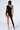 A person stands with their back to the camera, showcasing the ICONS ONLY STRETCH BODYSTOCKING in black, which features geometric cutouts along one leg and a single long sleeve. The stylish bodystocking reveals several tattoos on their upper and lower back. They complement the look with high-heeled shoes.