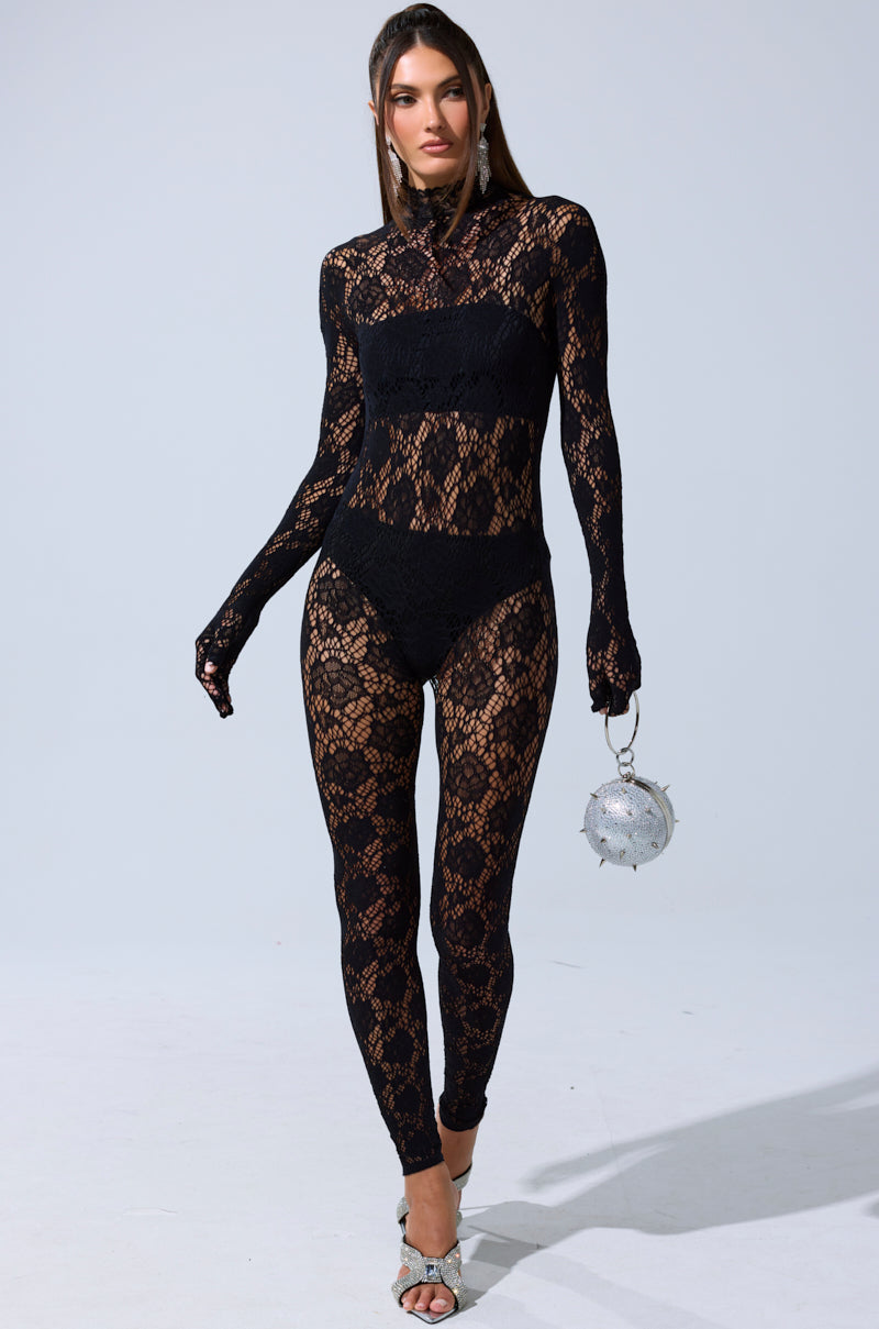A woman models the FEELIN SPICY STRETCH BODYSTOCKING—a sheer black lace long-sleeve piece with visible black undergarments—paired with a silver spiked purse and silver heels, posing against a plain light background.