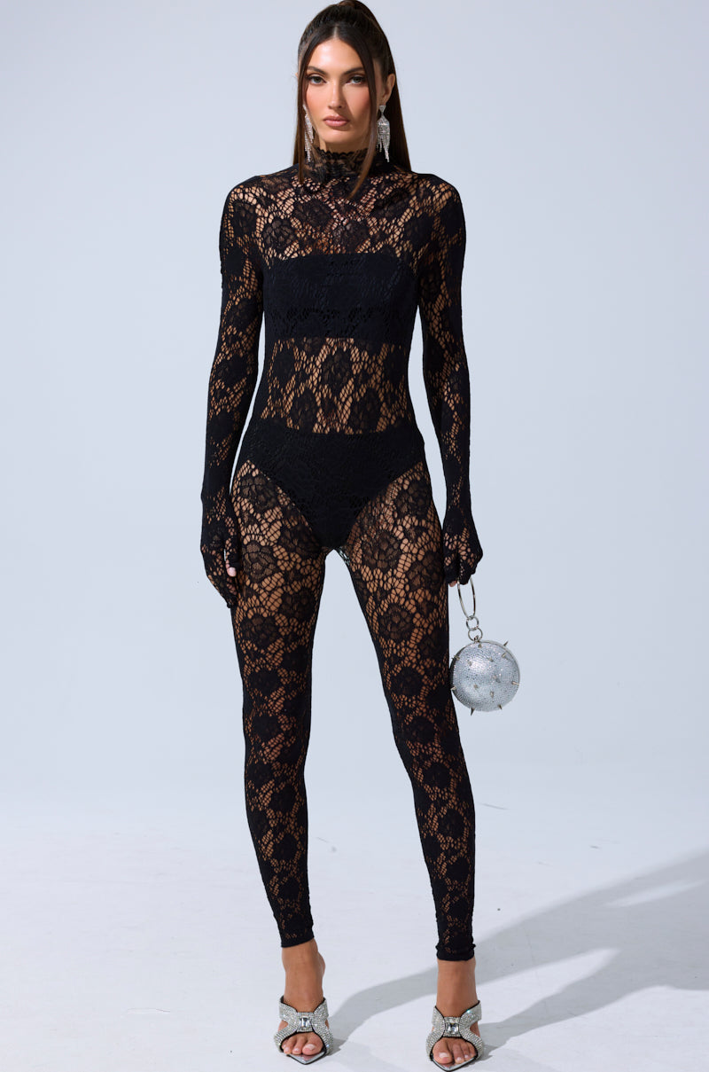 A woman faces forward in the FEELIN SPICY STRETCH BODYSTOCKING, a fitted black lace jumpsuit with long sleeves, high neck, and sheer details. She accessorizes with a small silver round purse and silver open-toe heels, with straight hair and subtle makeup.