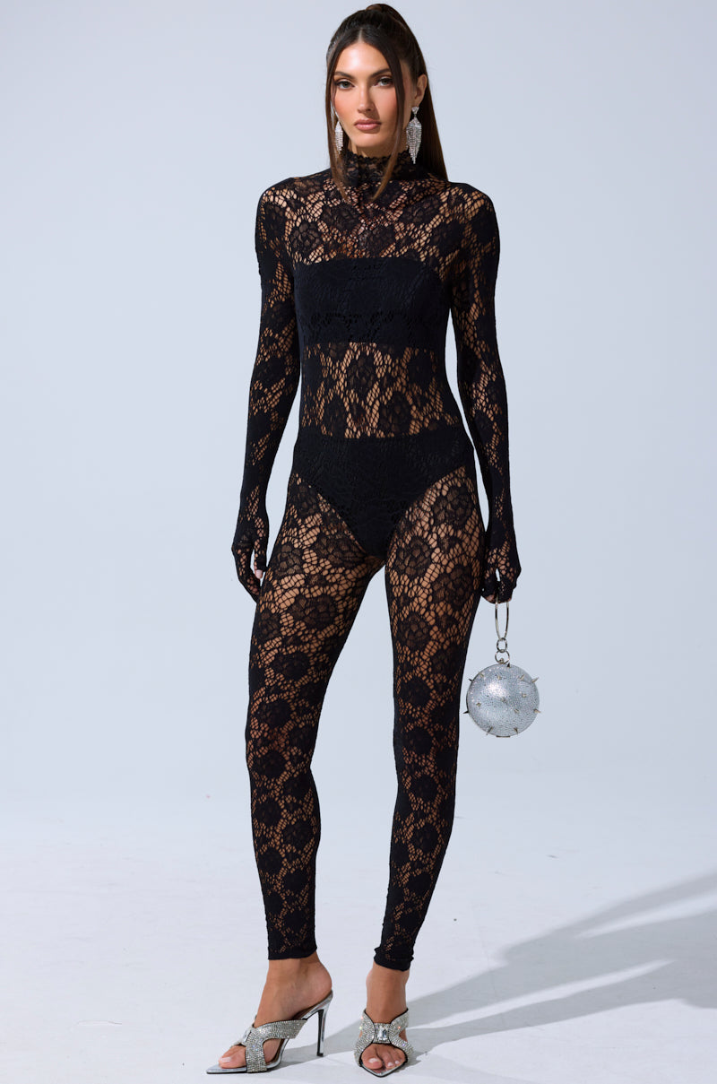 A woman faces forward wearing the FEELIN SPICY STRETCH BODYSTOCKING over black undergarments, paired with silver high-heeled sandals and a round silver purse. Her long hair is straight, and she accessorizes with visible earrings.