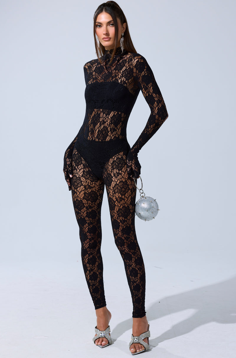 A woman stands against a plain background in the FEELIN SPICY STRETCH BODYSTOCKING—a black lace, long-sleeve, sheer bodysuit paired with black undergarments, silver heels, and a silver round handbag.