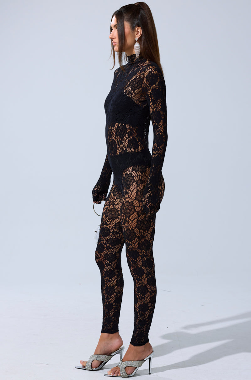 A woman stands in profile against a plain background, wearing the FEELIN SPICY STRETCH BODYSTOCKING—a sheer black lace jumpsuit with long sleeves and leggings. She accessorizes with statement earrings and silver high-heeled sandals.