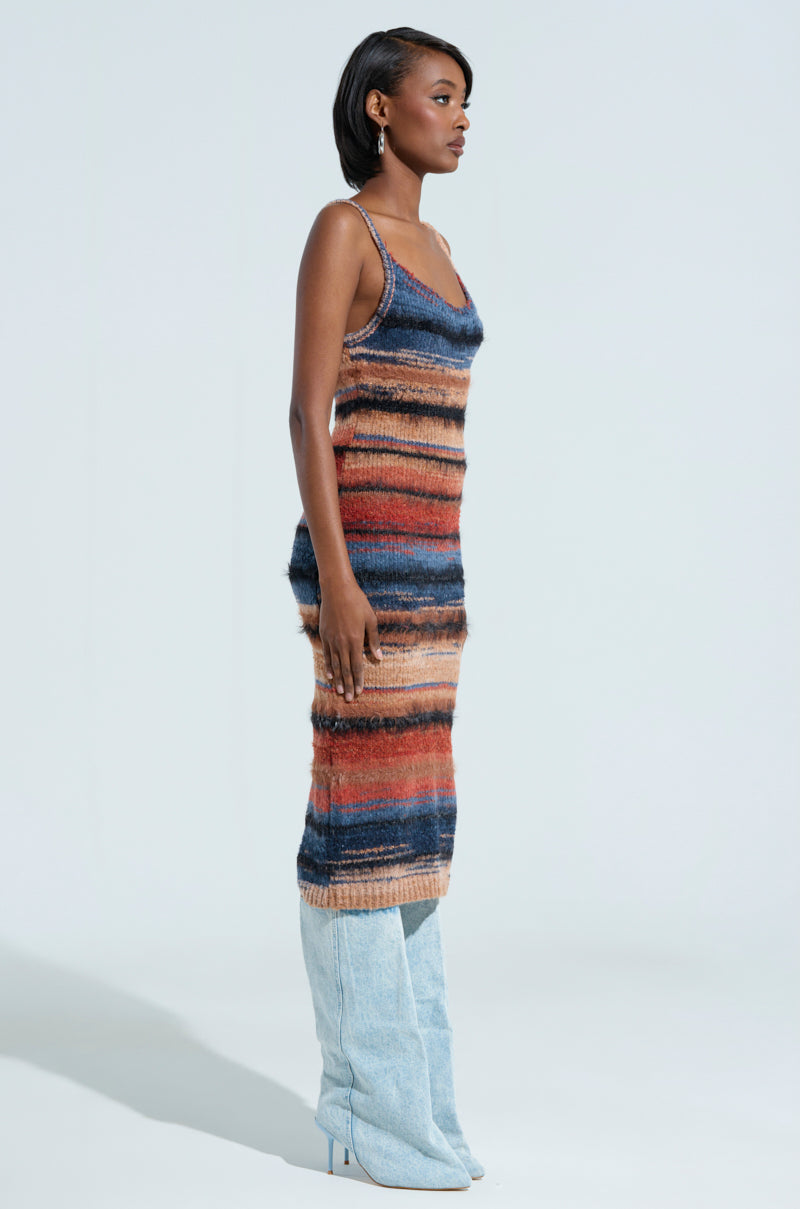 A person stands sideways, flaunting a curve-hugging silhouette in a vibrant knit dress—reminiscent of the UP ALL NIGHT MIDI DRESS—and light blue denim boots. Their short, dark hair creates an elegant contrast against the plain background.