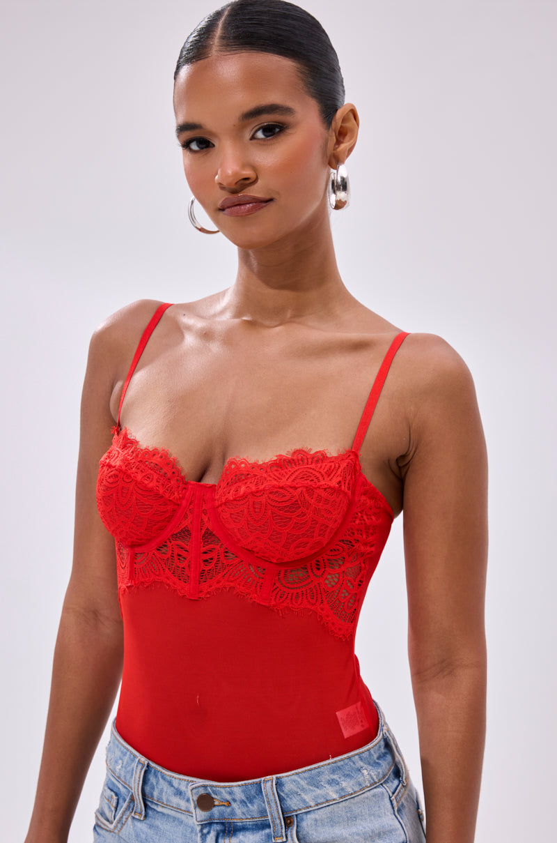 A woman confidently models the BURNING PASSION LACE BODYSUIT IN RED with blue jeans, her hair pulled back and hoop earrings on, standing against a plain light background.