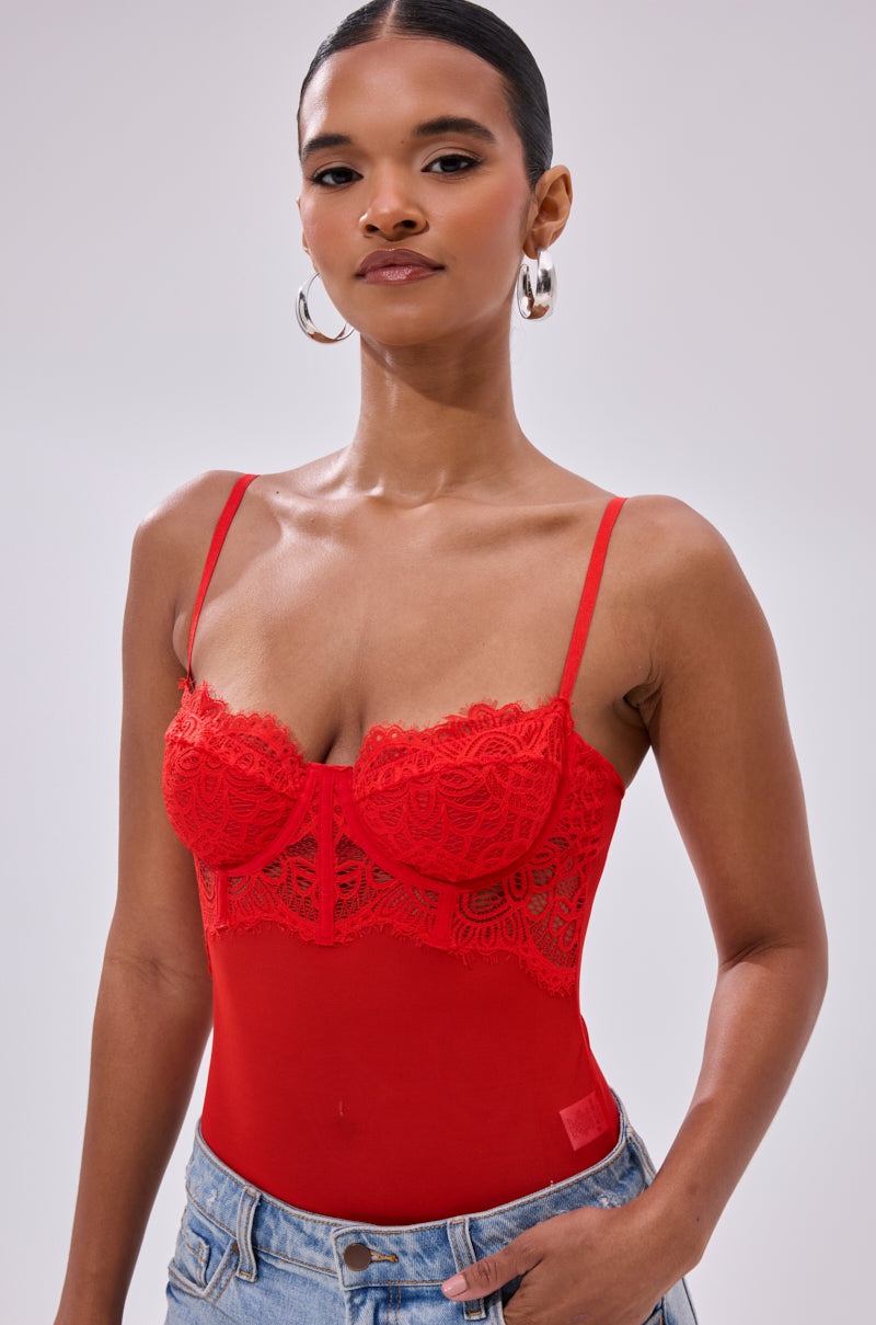 A woman wears the BURNING PASSION LACE BODYSUIT IN RED, a sheer mesh design with thin straps, paired with blue jeans. She stands against a plain light background, accessorized with hoop earrings and her hair pulled back.