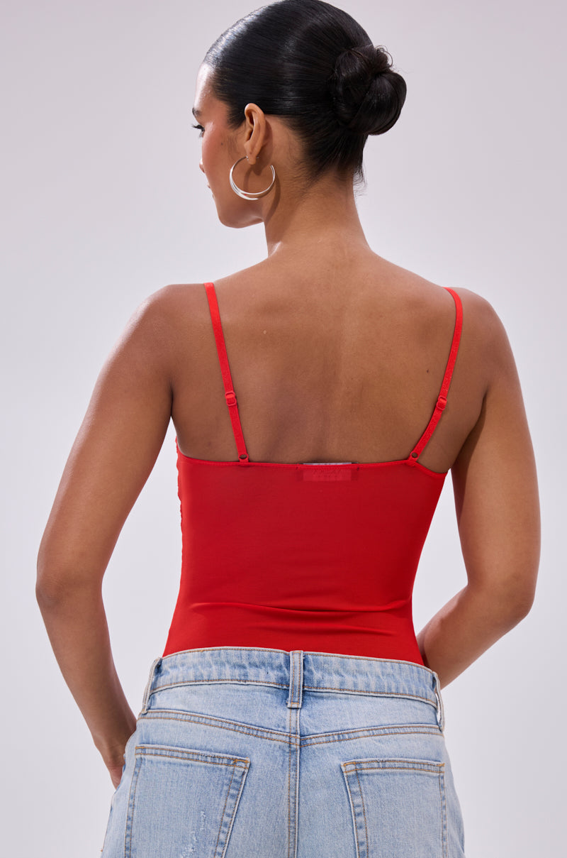 A woman with dark hair in a sleek bun models the BURNING PASSION LACE BODYSUIT IN RED, paired with light blue high-waisted jeans. She faces away to reveal the stylish back, resting one hand on her hip.