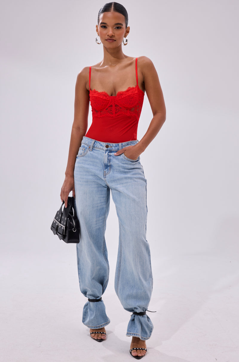 A woman wears the BURNING PASSION LACE BODYSUIT IN RED, light blue baggy jeans with tied hems, black strappy heels, hoop earrings, and holds a small black handbag against a plain light background.