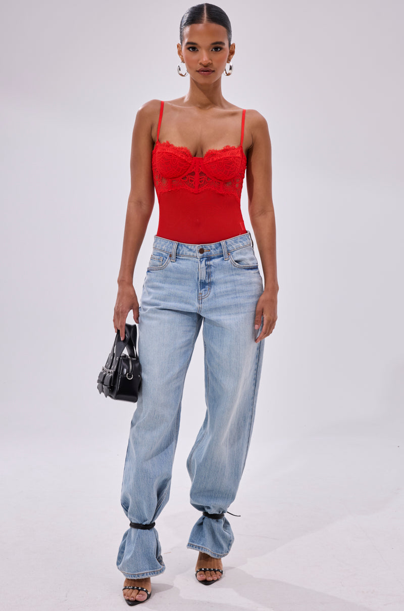 A woman faces forward in the BURNING PASSION LACE BODYSUIT IN RED, light blue baggy jeans tied at the ankles, black heeled sandals, gold hoop earrings, and holding a black handbag against a plain white background.