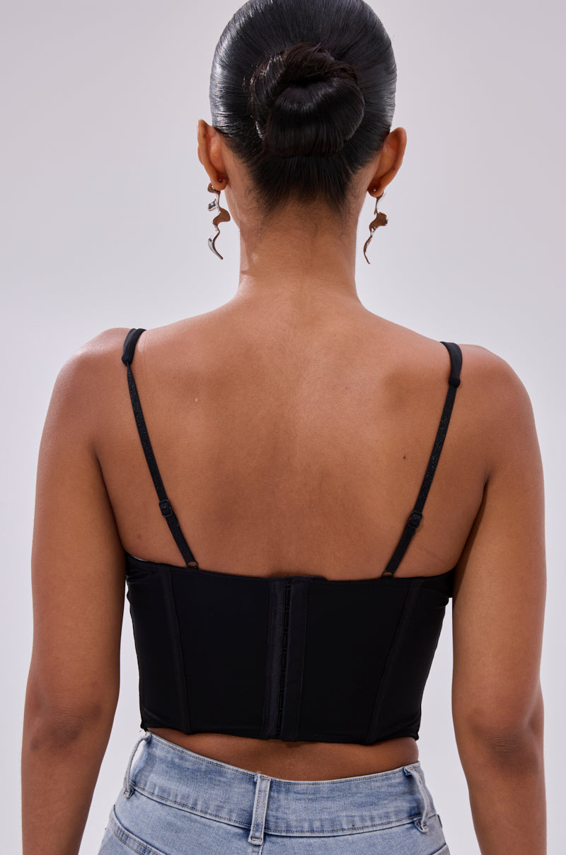 A woman with her hair in a neat bun is seen from the back wearing the FREE SPIRIT SLEEVELESS CORSET TOP IN BLACK, paired with light blue jeans and dangling abstract face-shaped earrings.