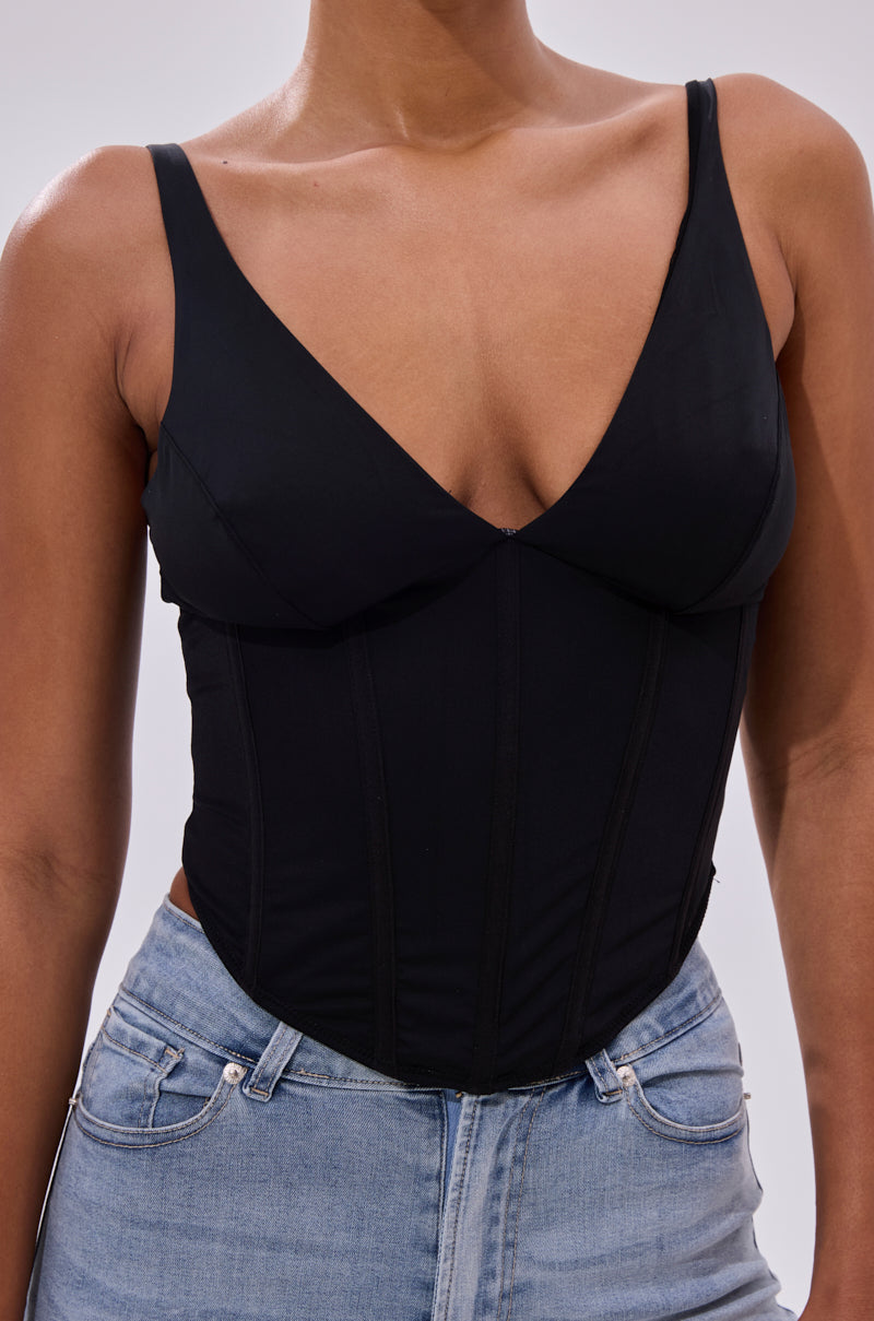 A person wears the FREE SPIRIT SLEEVELESS CORSET TOP IN BLACK, featuring a deep V neckline, paired with light blue jeans. The cropped corset top stands out against a plain, neutral background.