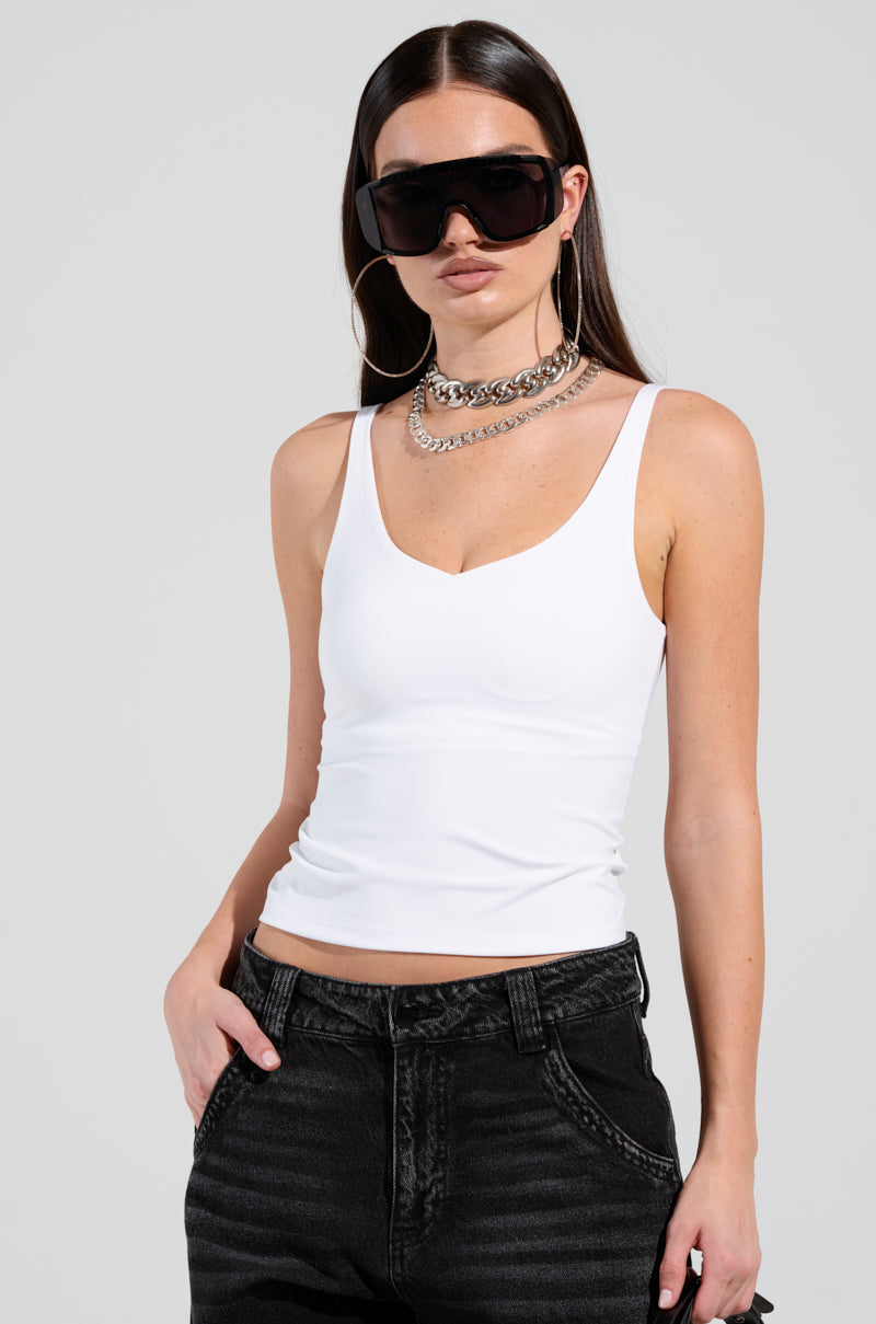 A person with long dark hair wears large black sunglasses, the JUMP AROUND TANK from AKIRA Label, and black jeans. They accessorize with hoop earrings and layered silver necklaces. The buttery soft fabrication of the tank complements their pose as they stand with one hand in their pocket against a plain background.