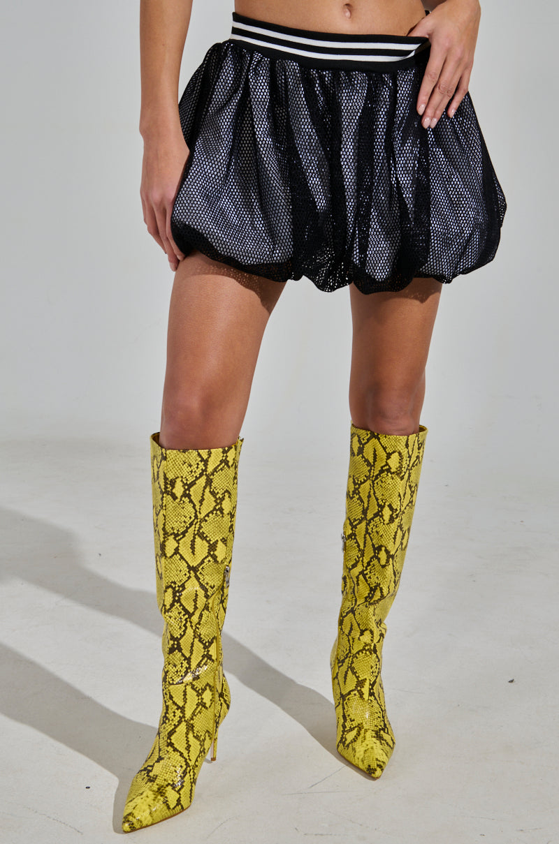 A person models a sporty-chic look featuring the PLAYERS ONLY JERSEY BUBBLE SKIRT, a black mesh design, paired with knee-high yellow snake-print boots. The sleek waistband and neutral gray background enhance this striking ensemble's lower half.