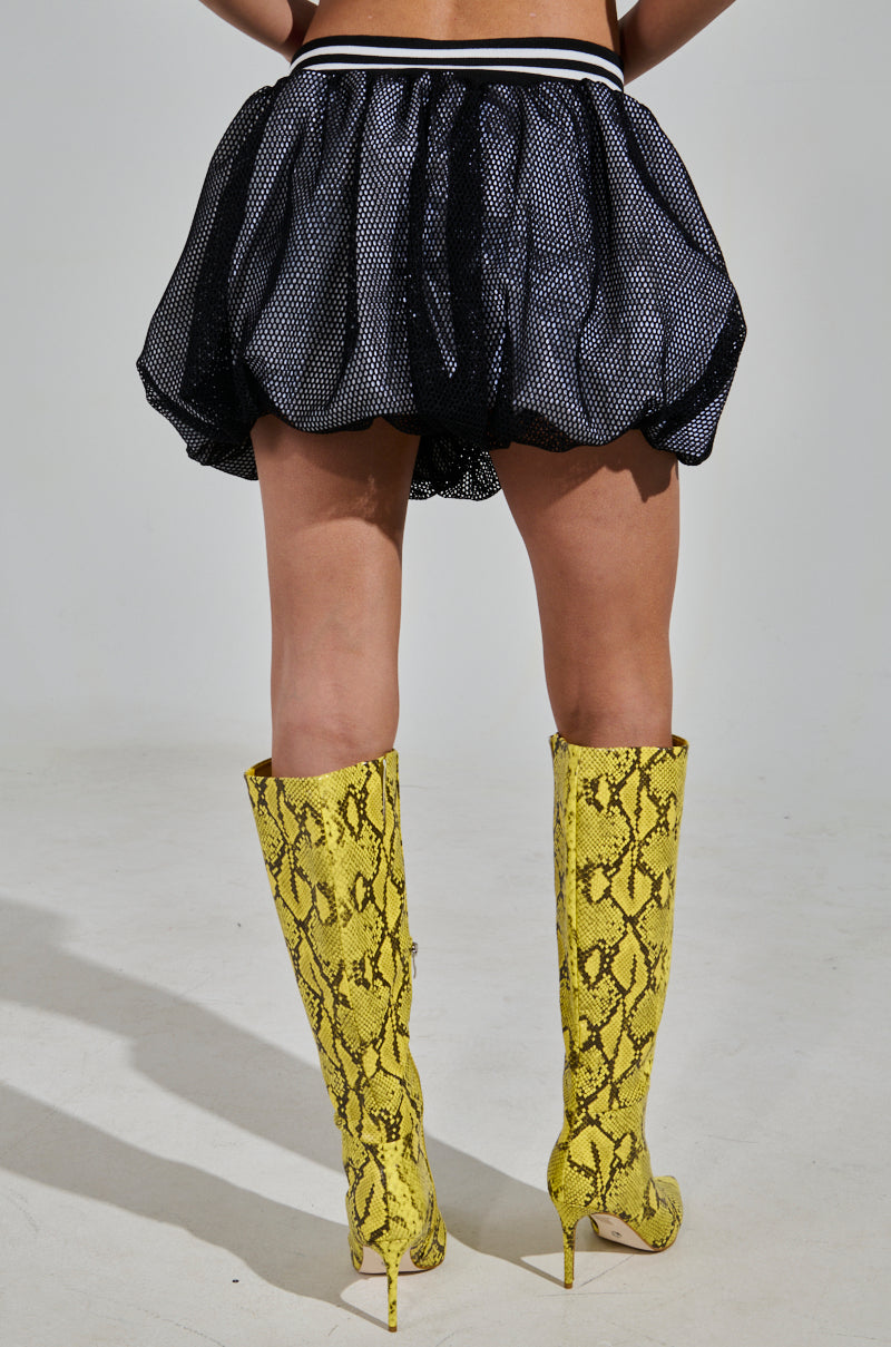 The image features a sporty-chic look with the PLAYERS ONLY JERSEY BUBBLE SKIRT, black mesh design, and yellow snake print knee-high boots with stiletto heels. Shot from behind, it highlights the flared skirt and tall boots.