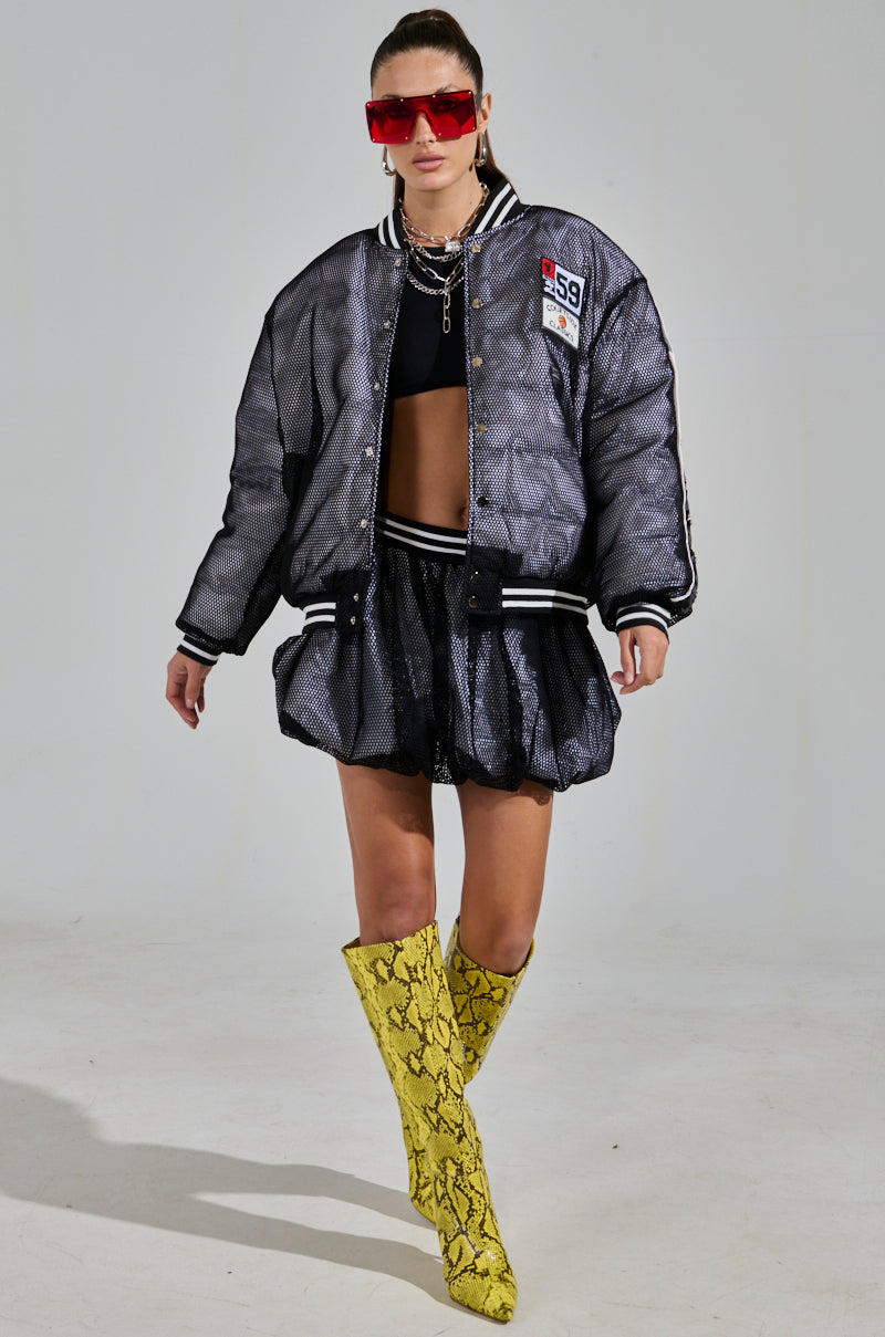 A person sports a sheer black bomber, black crop top, and the PLAYERS ONLY JERSEY BUBBLE SKIRT paired with bright yellow snake print boots for a sporty-chic vibe. Red sunglasses and a chunky necklace enhance the look against a plain gray backdrop, offering an unexpected twist.