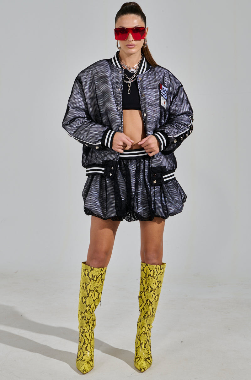 Wearing a metallic bomber jacket and black outfit, they pair yellow snake-patterned knee-high boots with the PLAYERS ONLY JERSEY BUBBLE SKIRT. Red sunglasses and layered necklaces add to their bold, sporty-chic style as they pose confidently against a plain background.