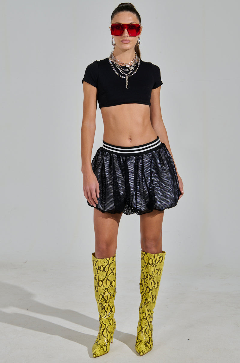 Sporty-chic style shines through in a black crop top and AKIRA Label's PLAYERS ONLY Jersey Bubble Skirt, paired with bold yellow snake-print knee-high boots. Red sunglasses and layered silver necklaces add flair against the plain gray backdrop.