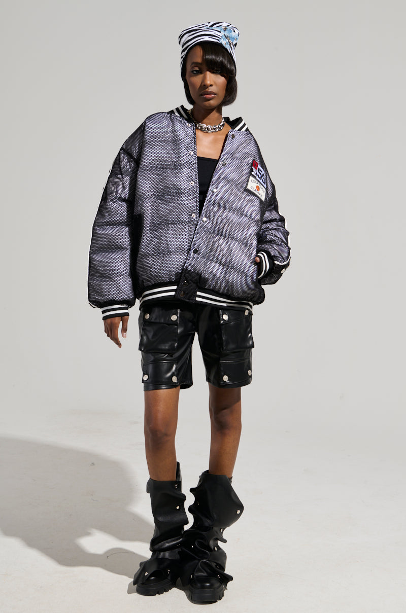 A person dressed in the PLAYERS ONLY JERSEY BOMBER, a large, shiny bomber with a checkered pattern and athletic patches, paired with black leather shorts featuring metal buttons and uniquely designed bulky black boots. They accessorize with a striped beanie and necklace against a plain, light-colored background.