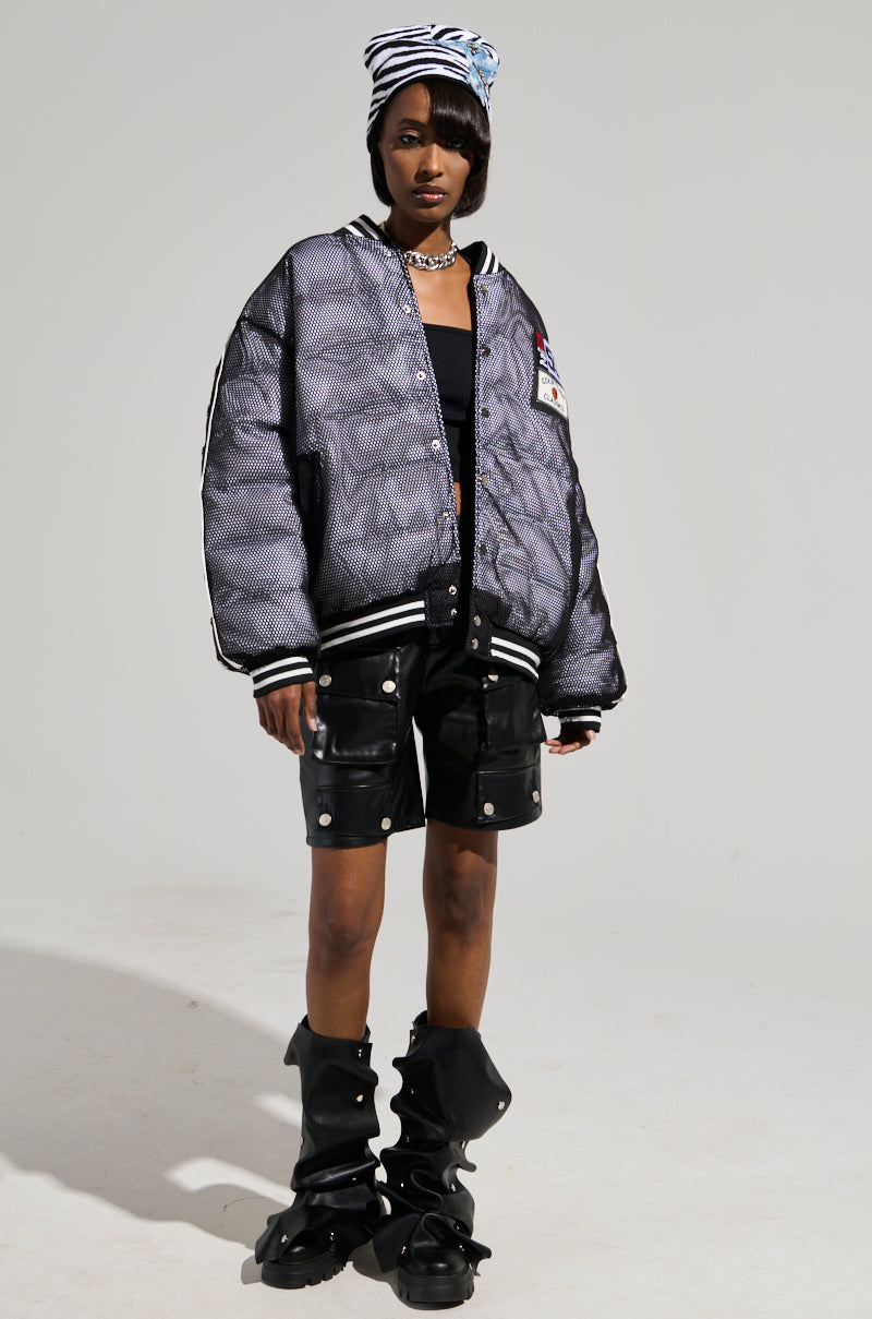 A person is wearing a PLAYERS ONLY JERSEY BOMBER jacket adorned with athletic patches, black shorts featuring silver buttons, and distinctive black shoes with bold designs. They also have a striped beanie and are posing against a plain background.