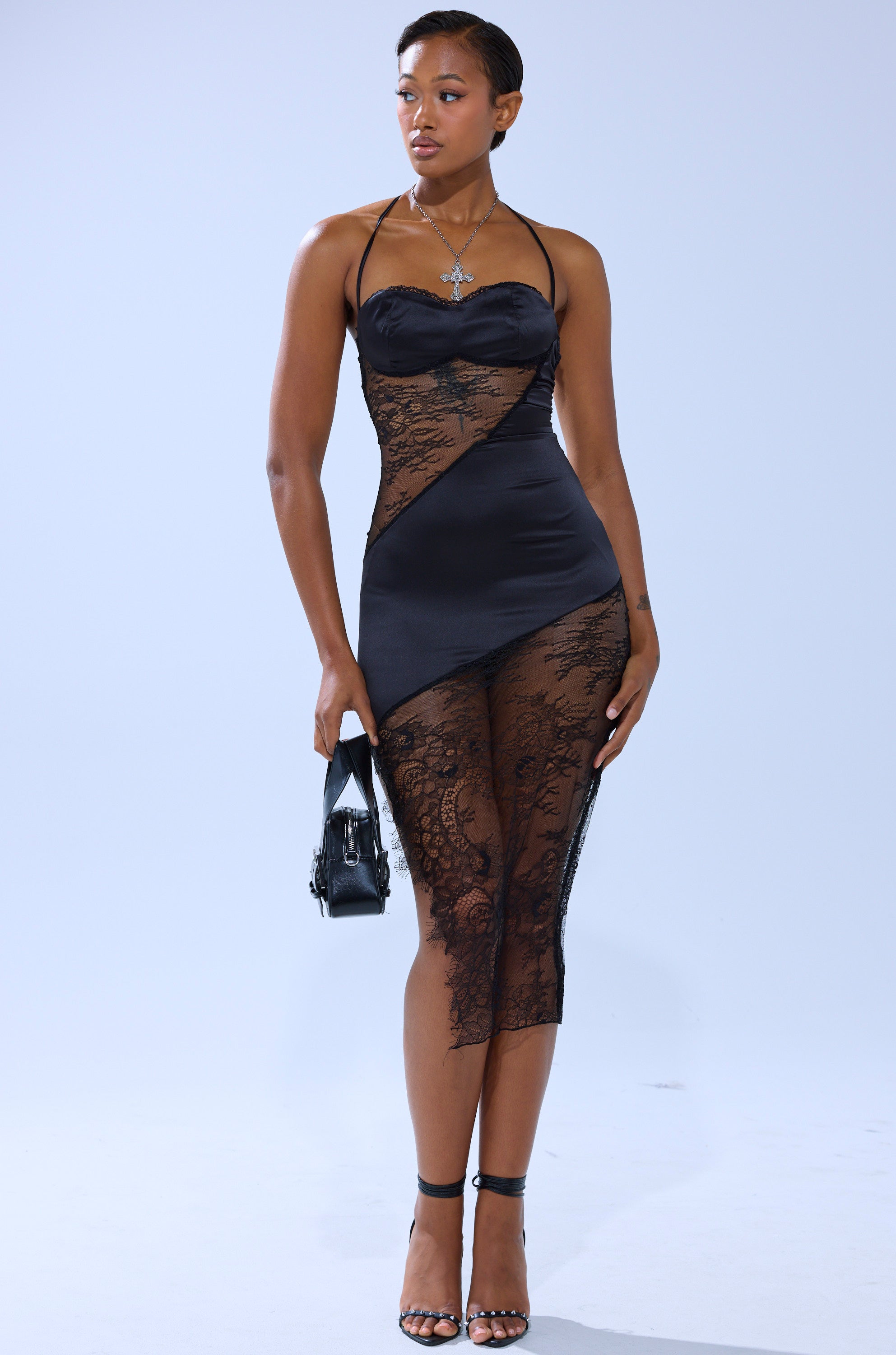 A woman with short hair stands against a plain light background, wearing the FEELIN MYSELF LACE CUTOUT MIDI DRESS, strappy heels, a necklace, earrings, and holding a small black purse.