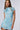 A woman with long, straight brown hair is wearing the LUANA MESH MINI DRESS, a light blue, short-sleeve dress adorned with a cherry blossom pattern. The dress features a ruffled hem and tie details near the shoulder. She is posing against a neutral background.