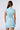 A person with long, straight hair is seen from behind wearing the LUANA MESH MINI DRESS — a short, light blue dress featuring a cherry blossom pattern, cap sleeves, and a ruffled hemline. They have one visible earring and are standing against a plain white background.